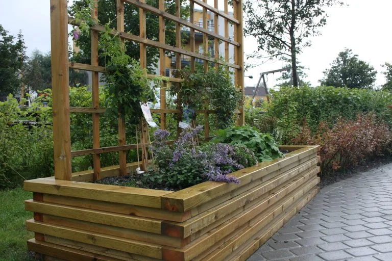 En stor trekasse med espalier inneholder grønne planter og blomster, plassert ved en gangsti omgitt av busker og trær.