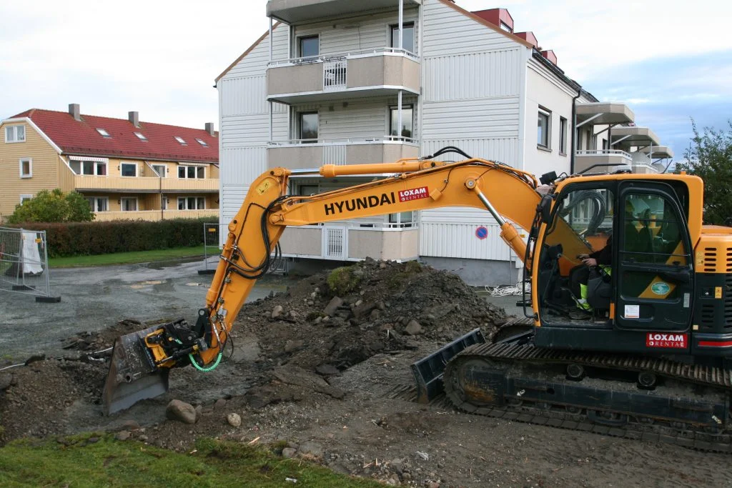 Gul gravemaskin fra Hyundai graver jord foran boligblokker