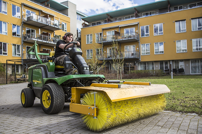 Grønn John Deere maskin med stor gul kost feier belegningsstein foran bygning