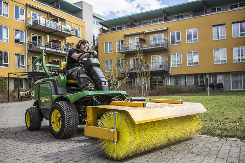 Grønn John Deere maskin med stor gul kost feier belegningsstein foran bygning