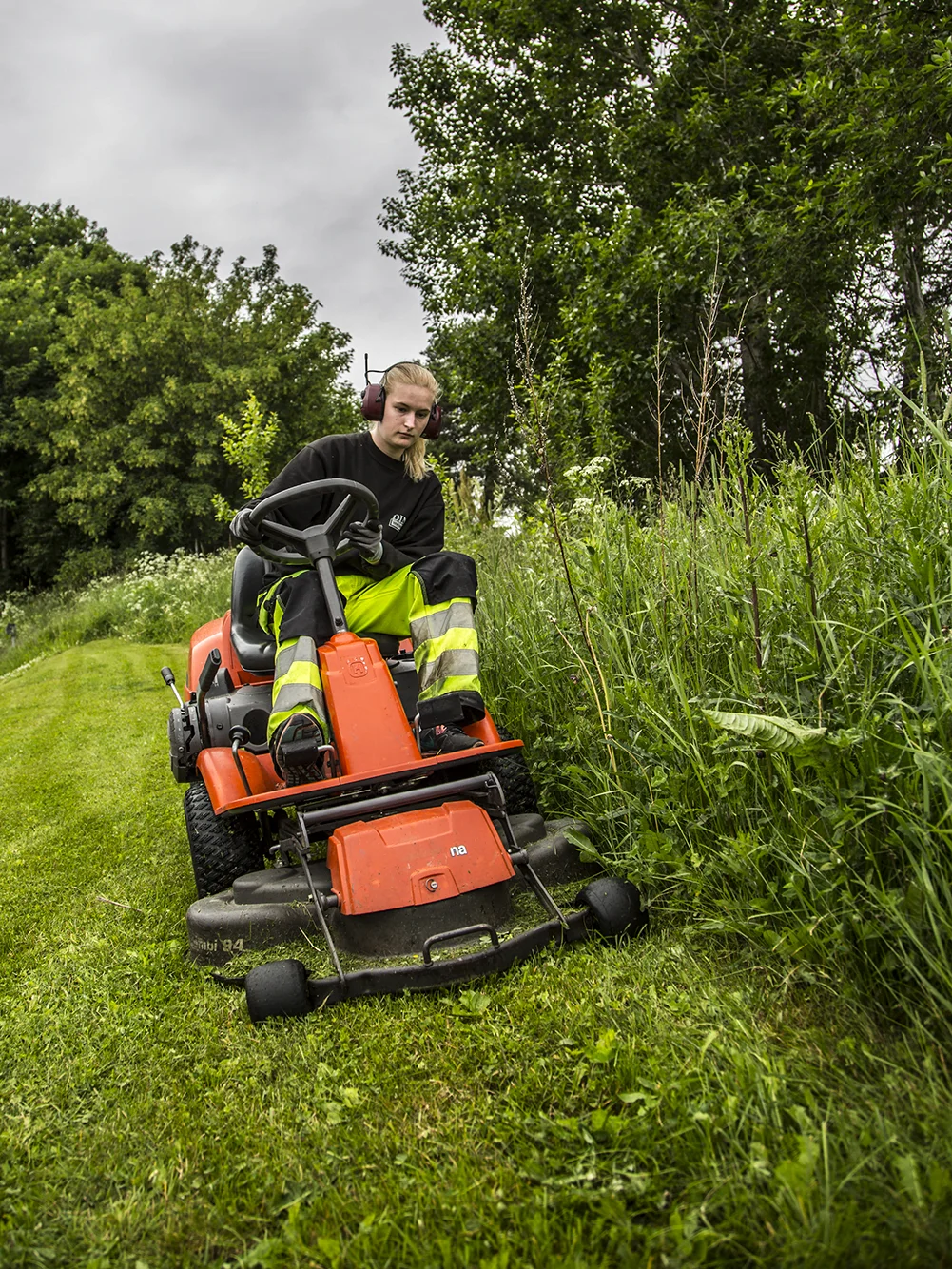 Person på oransje gressklipper klipper høyt gress ved grønn plen