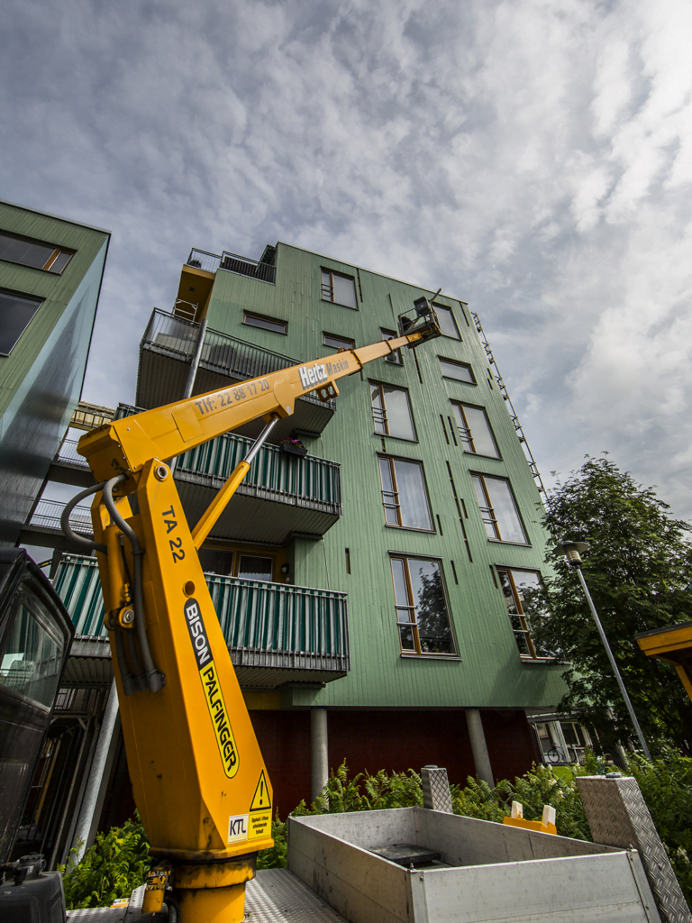 En gul lift med teleskoparm løfter en person opp til fasaden på et grønt leilighetsbygg for arbeid.