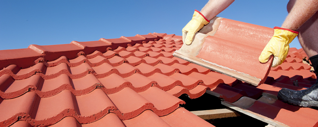En person med gule hansker legger en rød takstein på et tak under klar blå himmel.