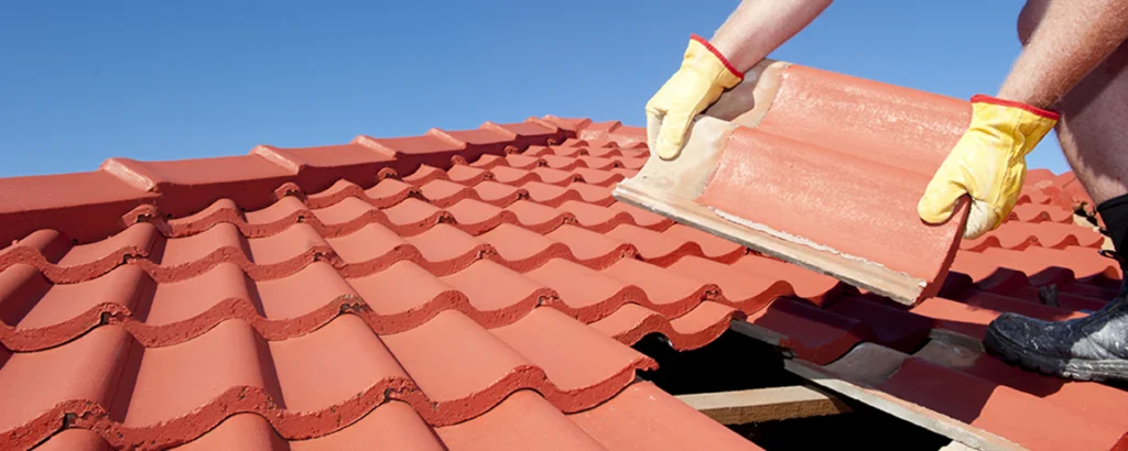 En person med gule hansker legger en rød takstein på et tak under klar blå himmel.