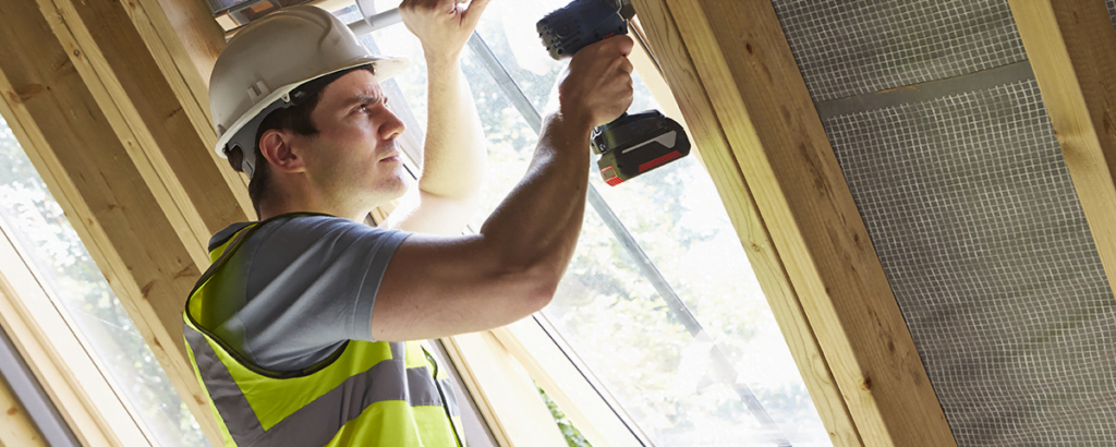 En person med hvit vernehjelm og gul refleksvest bruker en batteridrevet drill for å feste en ramme i en trekonstruksjon ved et vindu.