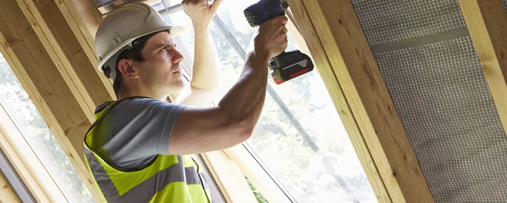 En person med hvit vernehjelm og gul refleksvest bruker en batteridrevet drill for å feste en ramme i en trekonstruksjon ved et vindu.