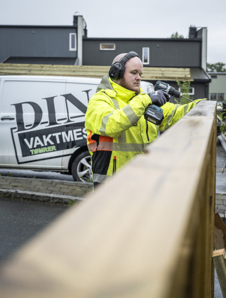 Person i gul refleksjakke fester skrue i treverk med batteridrill, varebil bak.
