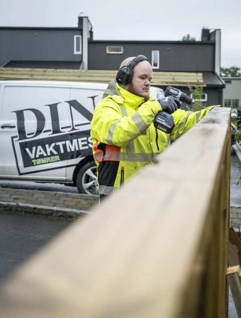 Person i gul refleksjakke fester skrue i treverk med batteridrill, varebil bak.
