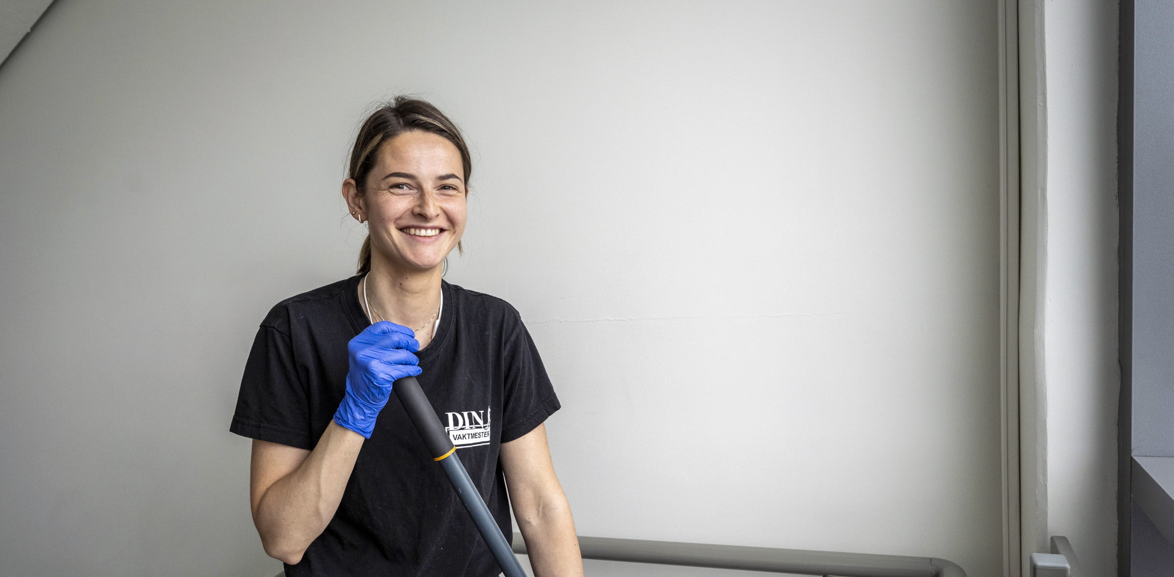 Person med blå hansker holder moppeskaft, iført svart t-skjorte med logo