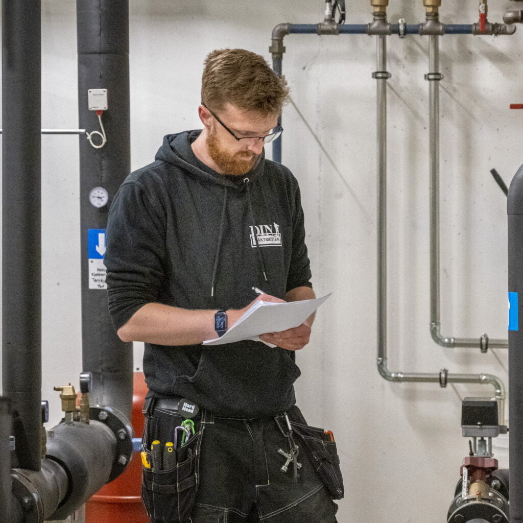 Person i svart hettegenser med verktøybelte skriver på ark i teknisk rom.