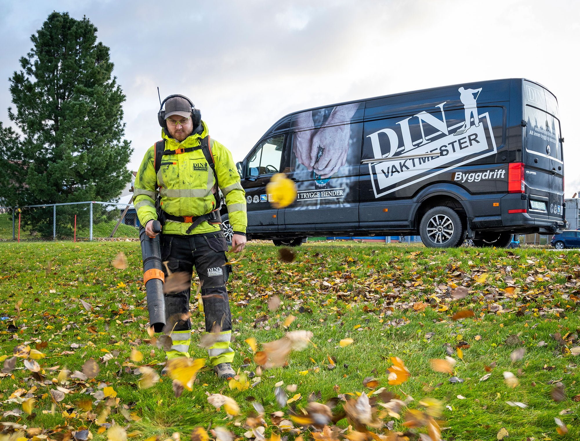 Person i gul refleksjakke bruker løvblåser på gress med høstløv, varebil bak
