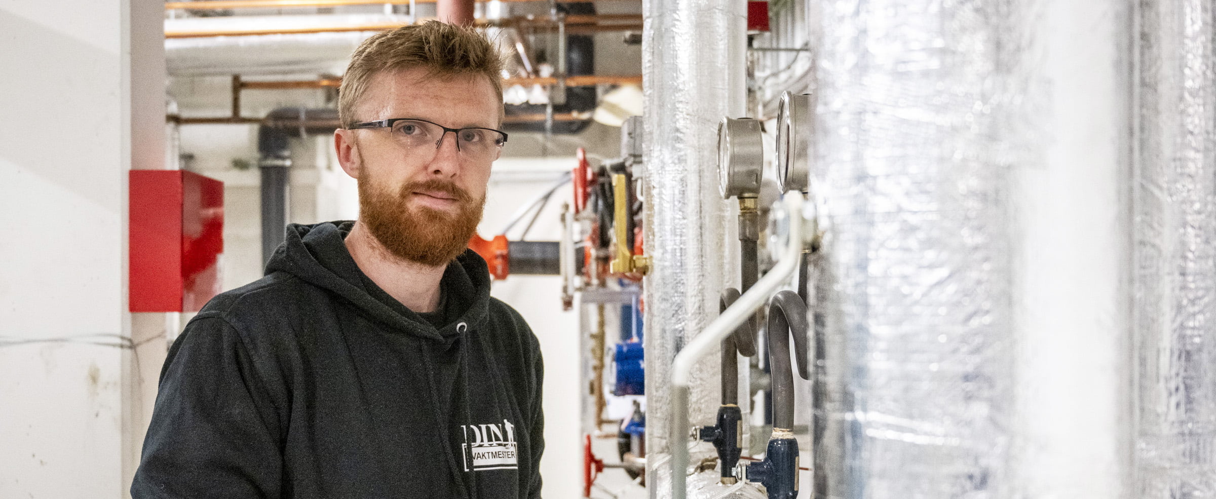 Person i mørk hettegenser jobber med rør og ventiler i teknisk rom.