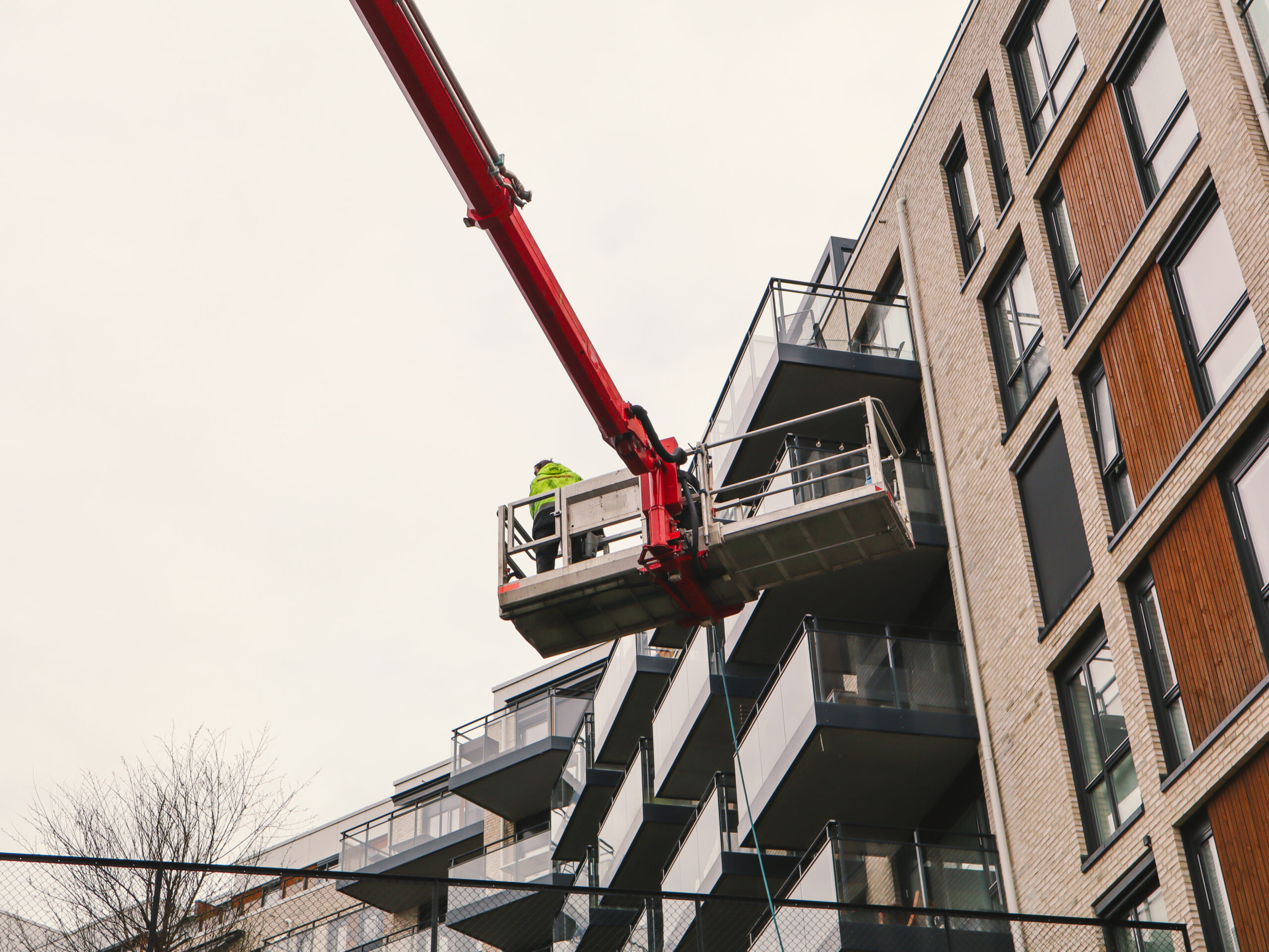 Person i gul refleksjakke står i liftkurv festet til rød kran ved balkonger.